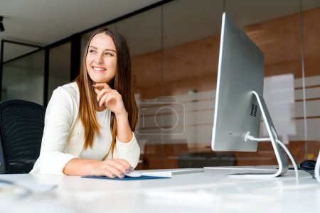 Téléchargez les photos : Une jeune femme d'affaires est capturée assise à son bureau, exprimant confiance et contentement alors qu'elle interagit avec un ordinateur dans un bureau élégant et contemporain. Souriante employée de bureau appréciant son travail - en image libre de droit