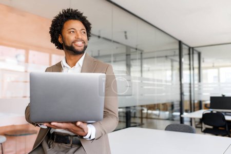 Téléchargez les photos : Homme d'affaires afro-américain en costume taupe tient son ordinateur portable et regarde au loin, reflétant la confiance et la vision dans un cadre de bureau moderne debout dans un espace de travail lumineux et ouvert - en image libre de droit