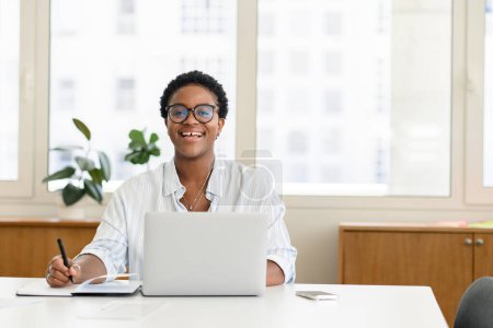 Téléchargez les photos : Étudiante africaine ravie, employée de bureau utilisant un ordinateur portable pour la recherche sur le Web, surfer. Femme utile en chemise décontractée prendre des notes dans un carnet, faire des recherches, regarder la caméra et rire - en image libre de droit