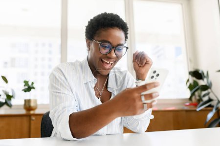 Photo for Overjoyed excited african american businesswoman looking at the phone screen, got job hiring, get good news or won in lottery. Victory concept, screaming yes - Royalty Free Image