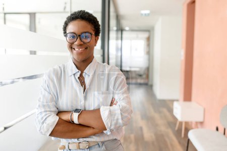 Téléchargez les photos : Portrait d'une élégante femme d'affaires afro-américaine debout dans le bureau moderne et posant contre la cloison de verre avec les bras croisés, regardant la caméra - en image libre de droit