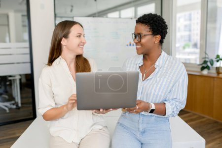 Photo for Friendly office atmosphere. Two female colleagues holding laptop and laughing looking at each other, having good communication inside team. Diverse colleagues in meeting room - Royalty Free Image