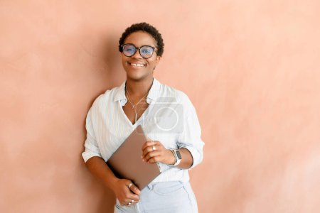 Photo for Young african woman freelancer stands in a modern office, posing confidently with a laptop in hand against wall. Inspired female employee looks at the camera cheerfully, feeling highly motivation - Royalty Free Image