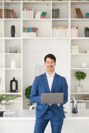 Foto de Un hombre de negocios hispano sonriente con un elegante traje azul se encuentra con un portátil en un ambiente de oficina elegante, encarnando la esencia de la elegancia empresarial moderna - Imagen libre de derechos