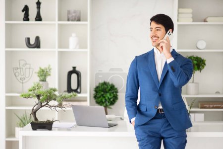 Photo for The Hispanic businessman enjoys a light-hearted moment during a phone call, his expression one of ease and positivity in a well-organized office space - Royalty Free Image