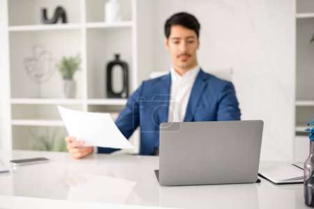 Photo for A professional Hispanic businessman in a blue blazer is focused on reading a document while sitting at a modern office desk, his laptop open in front of him. Male employee preparing annual report - Royalty Free Image