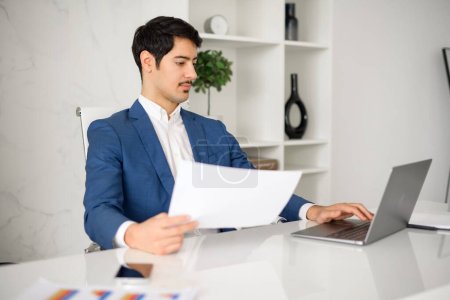 Photo for A thoughtful Hispanic businessman, wearing a blue jacket is reviewing a document while multitasking on his laptop in a contemporary office in a moment of critical thinking and business analysis - Royalty Free Image
