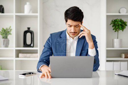 Téléchargez les photos : Homme d'affaires hispanique pensif regarde un ordinateur portable dans un espace de bureau, sa posture et l'expression reflétant la nature sérieuse de son travail. Un moment d'examen attentif ou de prise de décision difficile - en image libre de droit