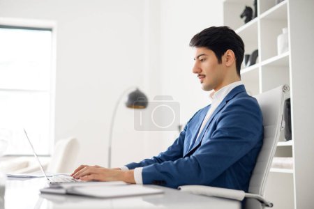 Photo for Professional Hispanic businessman in blue suit types attentively on a laptop in a bright modern office, showcasing productivity and digital proficiency, sitting in the office space, side view - Royalty Free Image