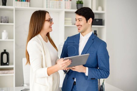 A friendly Hispanic businessman engages in a discussion with his female colleague over a tablet in a bright, modern office, suggesting effective communication and a positive work relationship