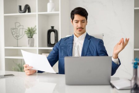 Photo for Hispanic businessman in a blue suit expressing confusion or concern while holding a document, seated at his office desk with a laptop, depicting a moment of business challenge or problem-solving - Royalty Free Image