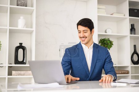 Photo for An attentive Hispanic businessman focuses on his laptop, surrounded by the minimalistic elegance of a modern office, demonstrating the dedication required in todays business environment - Royalty Free Image