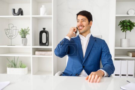 Photo for An upbeat Hispanic businessman talks on phone, holding a pleasant conversation, set in a modern office. Concept of effective communication and positive client interactions in a professional setting - Royalty Free Image