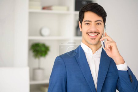 Photo for Charismatic Hispanic businessman in blue suit is engaging in conversation over smartphone, set against modern office, signaling blend of traditional business conduct and contemporary workspace design - Royalty Free Image