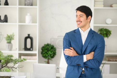 Photo for Young businessman smiling and standing in a relaxed pose with arms crossed in a bright office, communicate friendliness and a welcoming business atmosphere. A positive work environment concept - Royalty Free Image
