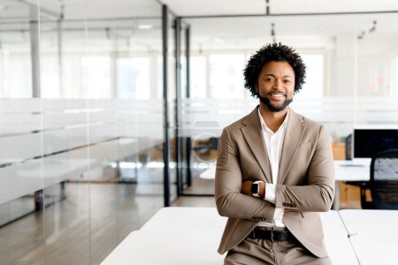 Téléchargez les photos : Homme d'affaires afro-américain élégant en costume marron clair pose les bras croisés dans un environnement de bureau moderne, respirant la confiance et l'accessibilité, propriétaire d'entreprise masculin, PDG, gestionnaire - en image libre de droit