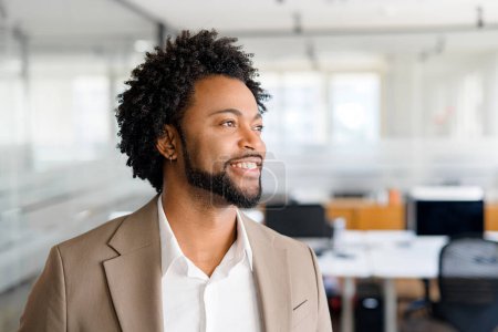 Foto de Hombre de negocios guapo y alegre en traje marrón mirando hacia otro lado con una sonrisa genuina, de pie en la oficina moderna, que representa la satisfacción laboral y el ambiente de trabajo positivo - Imagen libre de derechos