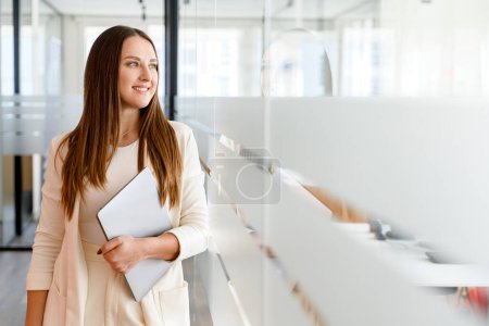 Foto de Joven profesional confiada en un traje de negocios ligero, sonriendo y llevando el portátil avanza a través del pasillo de la oficina, encarnando la naturaleza dinámica de los negocios modernos - Imagen libre de derechos