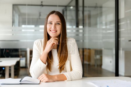 Foto de Una joven empresaria equilibrada se representa con una sonrisa agradable, sentada en su escritorio de la oficina contra un telón de fondo de tabiques de vidrio, sonriendo amigable empleada de la oficina mirando a la cámara - Imagen libre de derechos