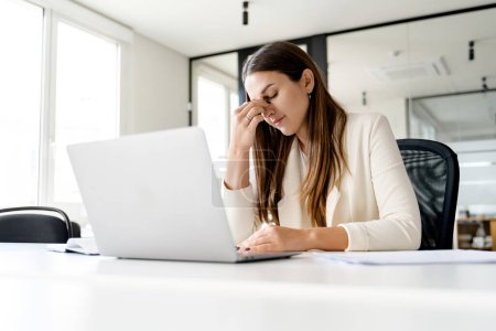 Una mujer de negocios preocupada pellizca el puente de su nariz mientras trabaja en su computadora portátil, una empleada de oficina que lidia con el estrés en el lugar de trabajo o reflexiona profundamente sobre un problema complejo