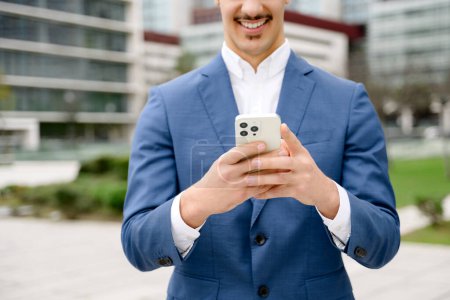 Photo for A close-up of cropped shot of Hispanic male entrepreneur using a smartphone, the device in hands of businessman, with the architecture of city on backdrop - Royalty Free Image