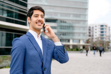 Photo for A young Hispanic businessman in a stylish blue suit speaks on the phone, with a bustling city life backdrop, embodying the concept of global connectivity and on-the-go business management. - Royalty Free Image