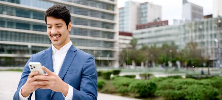 Photo for This panoramic shot captures a Hispanic entrepreneur engrossed in his smartphone, the wide city landscape behind him mirroring the expansive reach of modern business communication. - Royalty Free Image