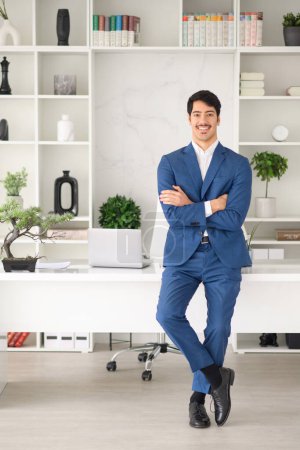 Photo for A confident Hispanic businessman stands with his arms crossed in a modern, bright office setting, wearing a sharp blue suit and a welcoming smile - Royalty Free Image