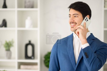 Foto de Amistoso y sonriente empresario hispano conversa en el teléfono inteligente, el diseño minimalista de la oficina acentuando su comportamiento profesional y enfoque moderno de los negocios - Imagen libre de derechos