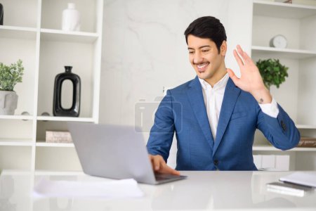 Photo for A jovial Hispanic businessman waves at his laptop screen during a video call, a representation of friendly remote communication and modern work practices. Virtual meeting concept - Royalty Free Image