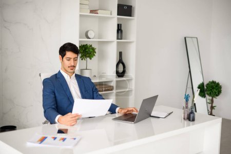 Photo for Young Hispanic businessman in formal wear looking intently at a paper document sitting with laptop in well-organized office space, represents a scene of daily corporate diligence and meticulous work - Royalty Free Image
