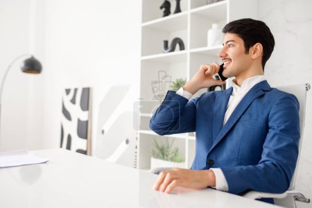 Téléchargez les photos : Un homme d'affaires hispanique confiant conversera sur son smartphone avec un sourire détendu, assis dans un bureau moderne et bien éclairé qui met l'accent sur la productivité et la connectivité.. - en image libre de droit