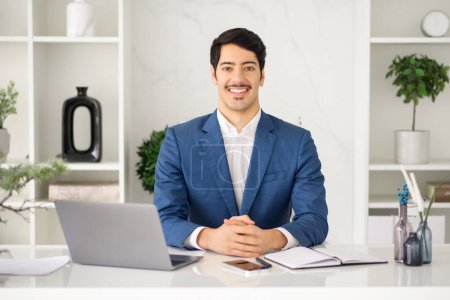 Ein lächelnder hispanischer Geschäftsmann sitzt an seinem Laptop in einem modernen Büro, das die Verkörperung moderner Professionalität und technologischer Integration im Unternehmenssektor ist.