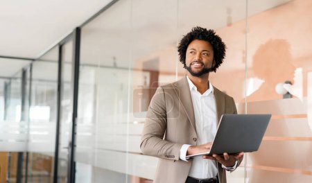 Homme d'affaires afro-américain regarde attentivement dans la distance tout en tenant son ordinateur portable, suggérant la planification stratégique et la contemplation pour les futures entreprises commerciales