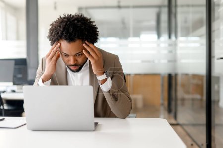 Photo for Stressed African-American businessman shows signs of concern as he intently studies his laptop screen in office. Concept of the challenges and pressures that come with professional responsibilities - Royalty Free Image