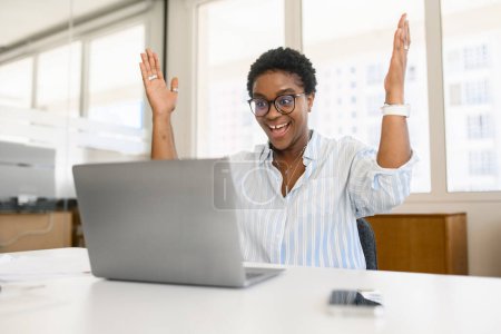 Foto de Feliz mujer afroamericana emocionada celebrando la victoria sentada en el co-trabajo en frente de la computadora portátil, afortunada mujer de negocios gritando sí, recibió buenas noticias, oferta de trabajo - Imagen libre de derechos