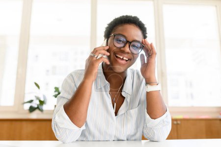 Foto de Alegre empleada afroamericana que usa una camisa informal inteligente que mantiene una conversación telefónica, mujer sentada en el escritorio de la oficina moderna, empresaria multirracial que habla en un teléfono inteligente y se ríe - Imagen libre de derechos