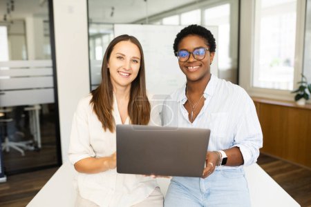 Foto de Dos felices y diversas colegas afroamericanas y caucásicas mujeres mirando a la cámara de pie en la oficina y sosteniendo el ordenador portátil, discutiendo juntos la estrategia - Imagen libre de derechos