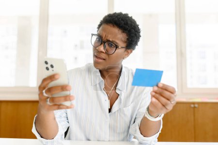 Téléchargez les photos : Jeune femme afro-américaine confus avec les cheveux courts tenant smartphone et carte de crédit, a une dette, employée de bureau ou pigiste ne reçoit pas de salaire, compte chèque solde avec visage triste - en image libre de droit