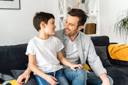 Photo for Happy together. Portrait of cheerful father and son sitting on the sofa in cozy living room, looking at each other and laughing, enjoying weekend at home - Royalty Free Image