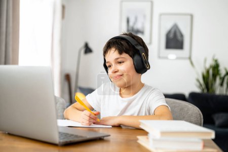 Photo for E-studying and homeschooling. Smiling schoolboy studying on the distance from home, using trendy laptop, preteen boy doing homework, writing tasks in the notebook, watching online lessons - Royalty Free Image