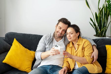 Foto de Alegre pareja pasa tiempo con un teléfono inteligente en casa, una esposa y un marido mira en la pantalla con sonrisas sentadas en el sofá en la acogedora sala de estar, viendo videos, desplazándose alimentar y reír - Imagen libre de derechos