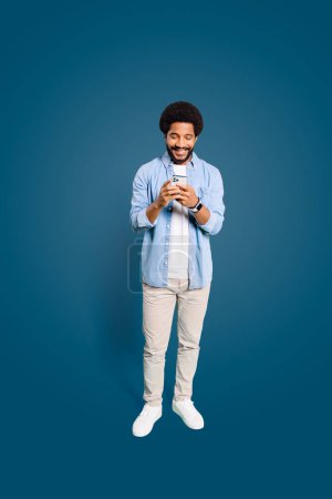 A cheerful young man with an afro hairstyle is holding a smartphone with both hands and smiling at the screen. The concept highlights modern connectivity and the joy of social interaction.