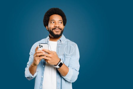 Photo for Young Brazilian man wearing a light blue denim shirt holds a smartphone and looks pensively to the side isolated on blue. Tech-savvy individual considering a new idea or message received on his phone. - Royalty Free Image