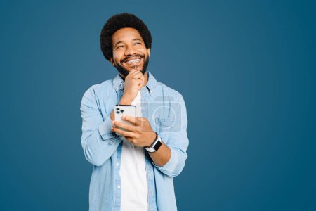 Téléchargez les photos : Un jeune Brésilien joyeux avec une coiffure afro, portant une chemise en denim, souriant et regardant de côté rêveur tout en tenant smartphone, recevoir de bonnes nouvelles ou profiter d'une conversation décontractée - en image libre de droit