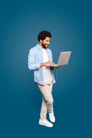 Photo for A young Brazilian man, male freelancer or student, standing and using a laptop, a picture of modern productivity on blue backdrop, representing the flexibility of remote work and digital nomadism - Royalty Free Image