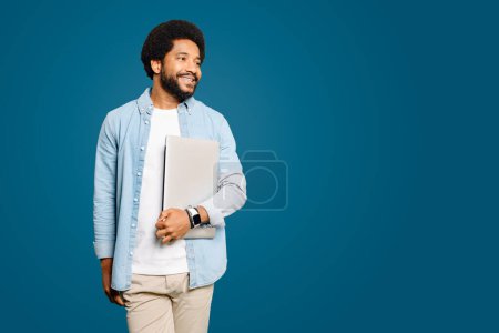 Photo for A smiling Brazilian young man, freelancer or student carrying a laptop standing isolated on blue, exuding confidence and readiness. Concept of readiness for business or learning opportunities. - Royalty Free Image