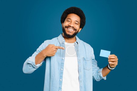 Foto de El hombre afroamericano apunta a una tarjeta de crédito con una sonrisa segura de pie aislado en azul, lo que sugiere la aprobación de un producto o servicio, o recomendar una herramienta financiera o aplicación. - Imagen libre de derechos
