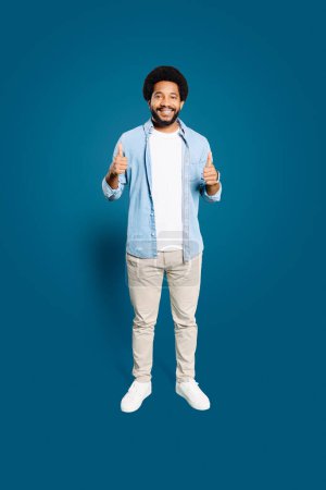 Photo for A cheerful man with frothy afro hair stands full-length, giving two enthusiastic thumbs up against a vibrant blue background, symbolizing positivity and approval, like-sign, recommend - Royalty Free Image