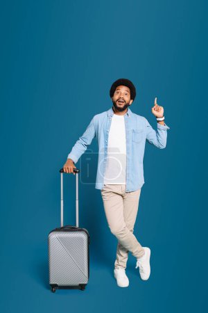 Photo for Young overjoyed man with suitcase lifts a finger as if having a bright idea about his travel plans, his cheerful demeanor suggesting an enjoyable journey ahead, full length photo isolated on blue - Royalty Free Image